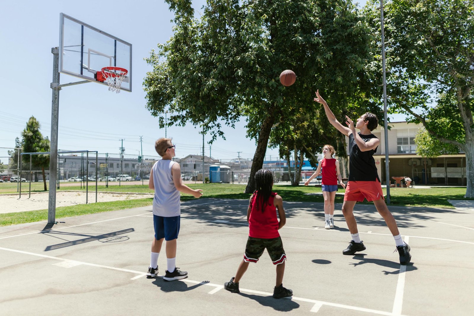 Group of children playing basketball on a sunny outdoor court with trees around.